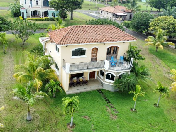 An aerial view of a Mediterranean-style house with a red-tiled roof, balcony, and lush greenery with palm trees surrounding it.