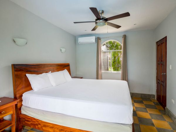 A clean bedroom with a wooden bed, white sheets, ceiling fan, window with curtains, wall-mounted air conditioner, and bedside tables.