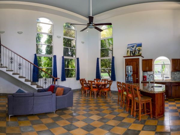 The image shows a spacious open-concept living and dining area with high ceilings, a staircase, and large windows with blue curtains, and a kitchen island.