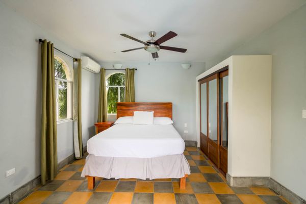 A bedroom with a bed, two windows with curtains, a ceiling fan, air conditioning, a wooden wardrobe with mirrors, tile flooring, and neutral walls.