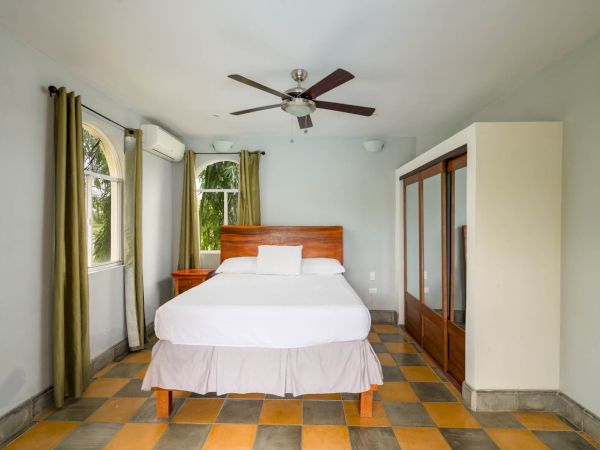A bedroom with a bed, two windows with curtains, a ceiling fan, air conditioning, a wooden wardrobe with mirrors, tile flooring, and neutral walls.