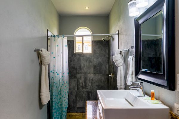 A bathroom with a sink, granite countertop, large mirror with lights, shower curtain, and a window above the shower area.