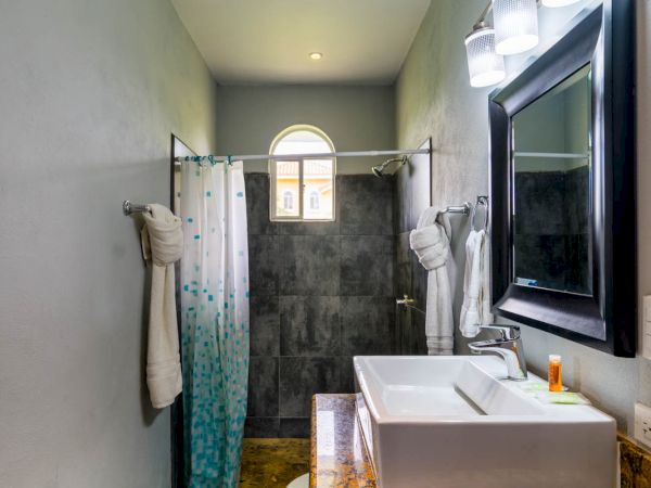 A bathroom with a sink, granite countertop, large mirror with lights, shower curtain, and a window above the shower area.