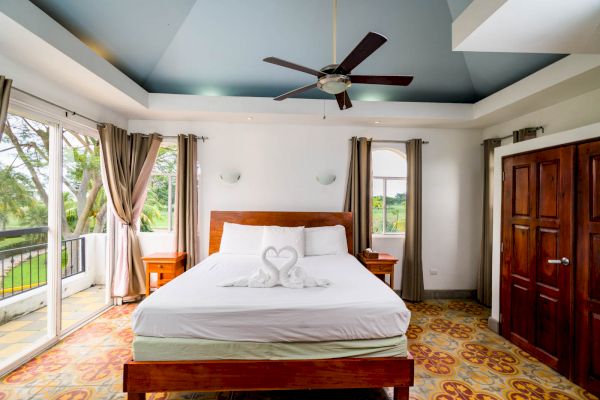 A bright bedroom has a bed with white linens and towel swans, a ceiling fan, wooden furniture, large windows, and an open view.