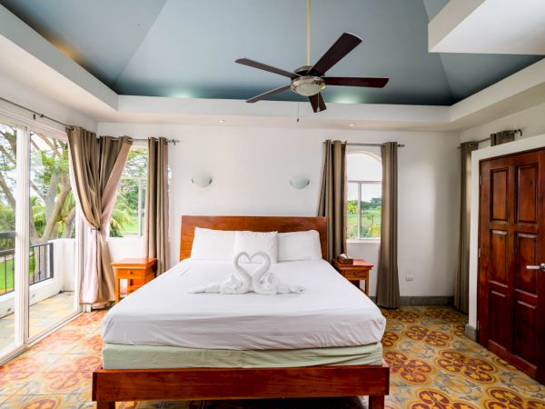 A bright bedroom has a bed with white linens and towel swans, a ceiling fan, wooden furniture, large windows, and an open view.