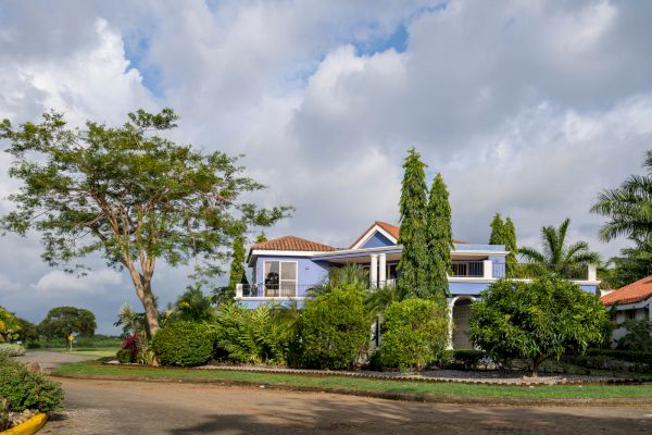 A two-story house with a red-tiled roof is surrounded by lush greenery and tall trees under a cloudy sky, located on a quiet street corner.