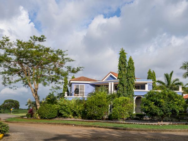 A two-story house with a red-tiled roof is surrounded by lush greenery and tall trees under a cloudy sky, located on a quiet street corner.