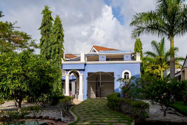 A two-story blue house with a red-tiled roof, surrounded by trees and a landscaped garden with a stone walkway leading to the entrance.