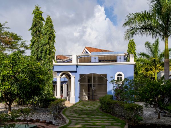 A two-story blue house with a red-tiled roof, surrounded by trees and a landscaped garden with a stone walkway leading to the entrance.