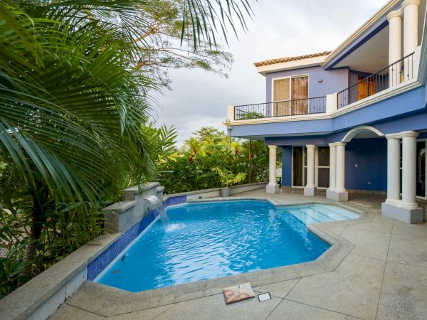 This image shows a luxurious home with a blue facade, featuring a private swimming pool surrounded by tropical plants and a spacious patio.
