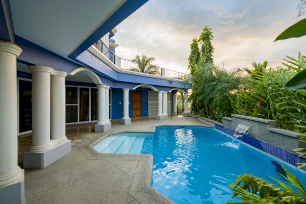 A luxurious outdoor area features a blue-tiled swimming pool, surrounded by a covered patio, white columns, tropical plants, and a cascading water feature.