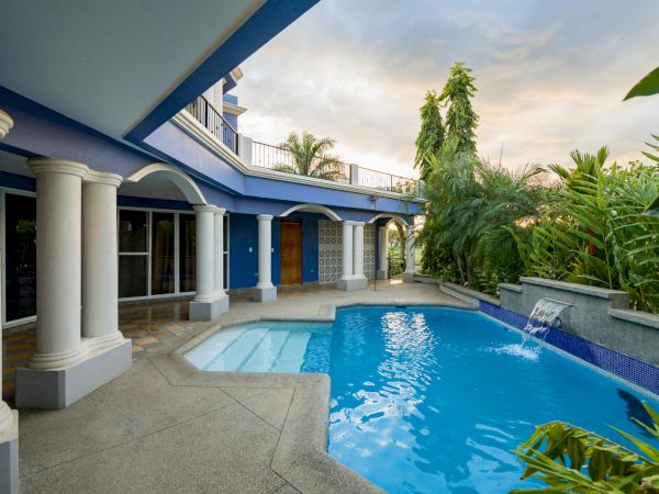 A luxurious outdoor area features a blue-tiled swimming pool, surrounded by a covered patio, white columns, tropical plants, and a cascading water feature.