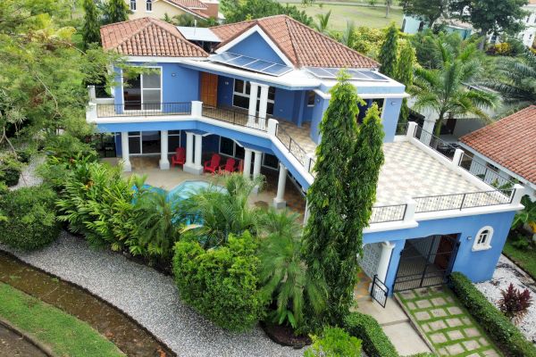 The image shows a two-story house with a blue exterior, surrounded by lush greenery and a gravel walkway, featuring a pool and patio area.