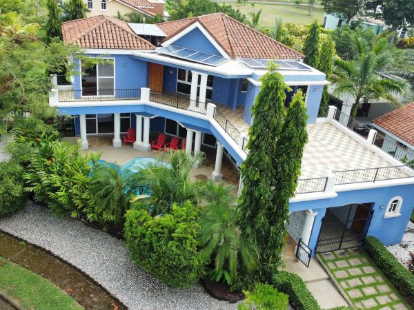 The image shows a two-story house with a blue exterior, surrounded by lush greenery and a gravel walkway, featuring a pool and patio area.