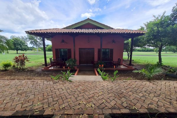 The image shows a small, one-story house with a brown roof, brick pathway, plants in front, surrounded by greenery, and a partly cloudy sky.