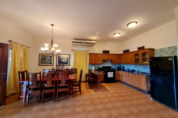 The image shows a spacious kitchen and dining area with wooden cabinets, a dining table, chairs, appliances, and tiled floors under soft lighting.