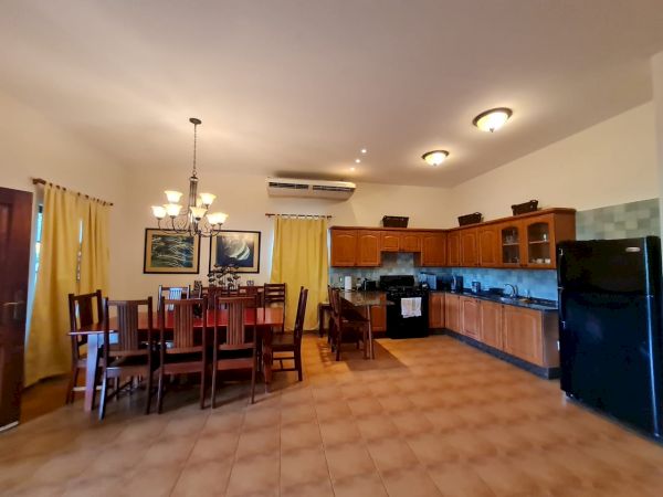 The image shows a spacious kitchen and dining area with wooden cabinets, a dining table, chairs, appliances, and tiled floors under soft lighting.