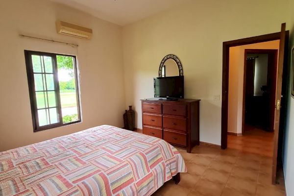 A bedroom with a bed, striped bedding, ceiling fan, window, dresser with a TV on top, and an open door leading to another room.