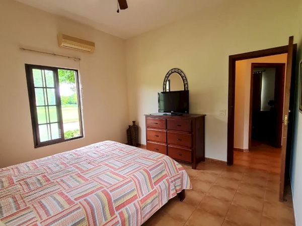 A bedroom with a bed, striped bedding, ceiling fan, window, dresser with a TV on top, and an open door leading to another room.