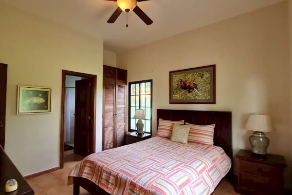 A cozy bedroom with a ceiling fan, double bed with striped bedding, bedside tables with lamps, and paintings on the walls, leading to a balcony.