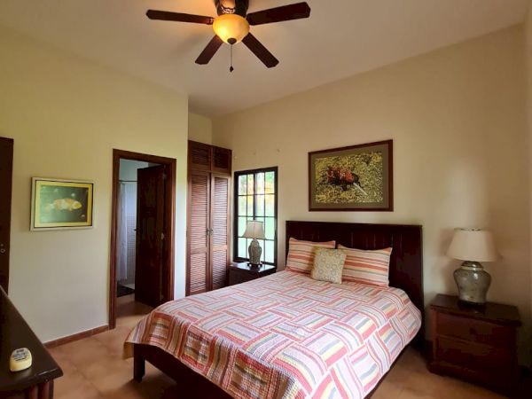 A cozy bedroom with a ceiling fan, double bed with striped bedding, bedside tables with lamps, and paintings on the walls, leading to a balcony.