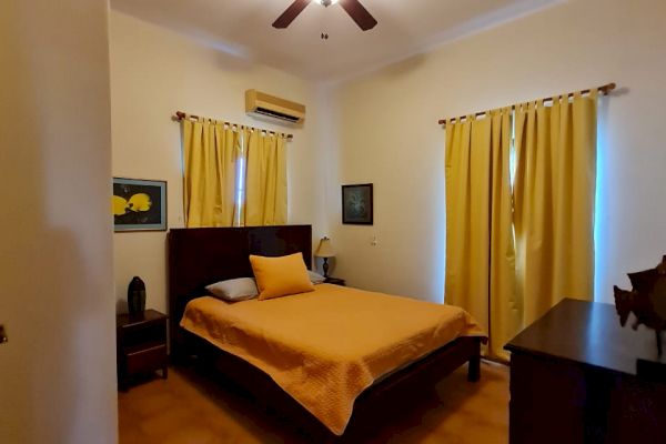 A bedroom featuring a bed with yellow bedding, matching yellow curtains, a ceiling fan, and a dresser. The room has a warm, cozy ambiance.