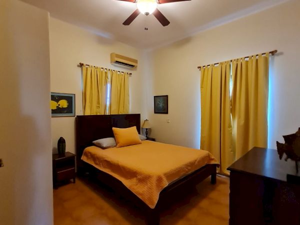 A bedroom featuring a bed with yellow bedding, matching yellow curtains, a ceiling fan, and a dresser. The room has a warm, cozy ambiance.