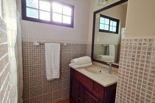 This image shows a small bathroom with a wooden vanity, sink, mirror, towel rack, and window. The walls are tiled, and there is a mat on the floor.