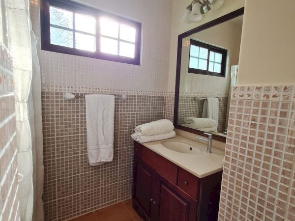 This image shows a small bathroom with a wooden vanity, sink, mirror, towel rack, and window. The walls are tiled, and there is a mat on the floor.