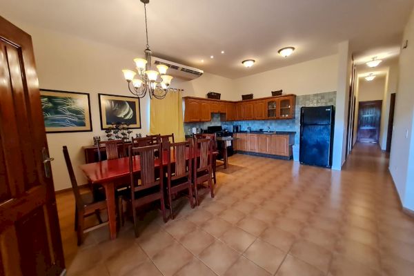 This image shows a dining area with a wooden dining table and chairs, a chandelier, and a spacious kitchen in the background with wooden cabinets.