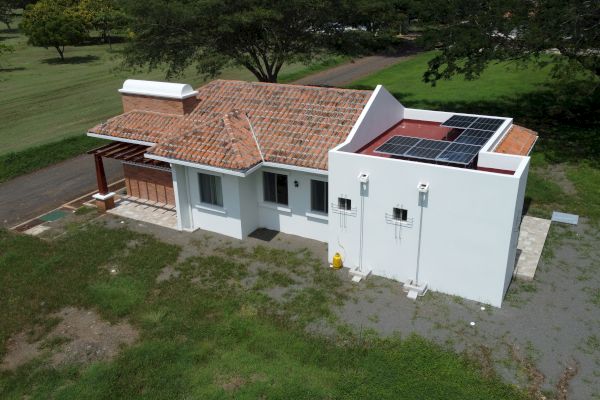 An aerial view of a small white house with a tiled roof, solar panels on the roof, and surrounded by grass and trees.