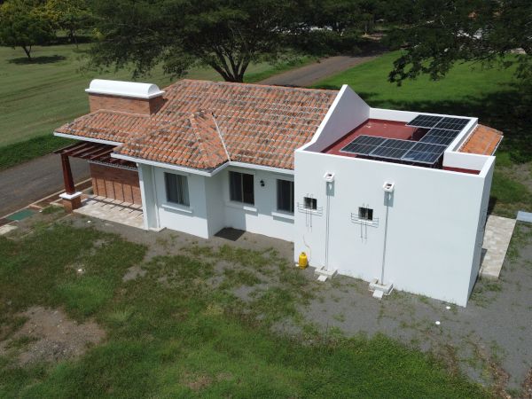 An aerial view of a small white house with a tiled roof, solar panels on the roof, and surrounded by grass and trees.