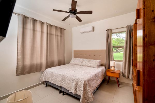 A neatly arranged bedroom with a bed, bedside table, ceiling fan, curtains, and a window showing a green view outside.