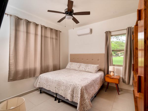 A neatly arranged bedroom with a bed, bedside table, ceiling fan, curtains, and a window showing a green view outside.