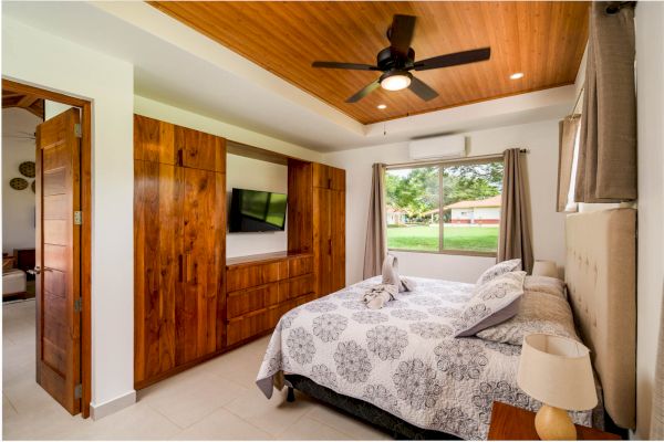 A bedroom with a bed, wooden wardrobe with a TV, ceiling fan, nightstand, and a large window with an outdoor view.