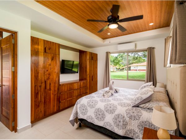 A bedroom with a bed, wooden wardrobe with a TV, ceiling fan, nightstand, and a large window with an outdoor view.