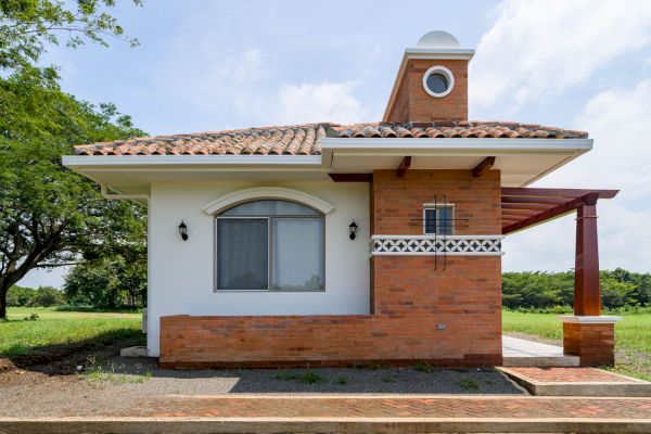 A small, single-story house with a combination of white and brick walls, surrounded by green grass and trees, under a clear sky ending the sentence.