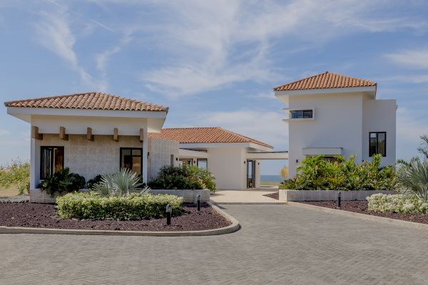 The image shows a modern villa complex with two buildings, paved pathways, landscaped gardens, and a clear blue sky.