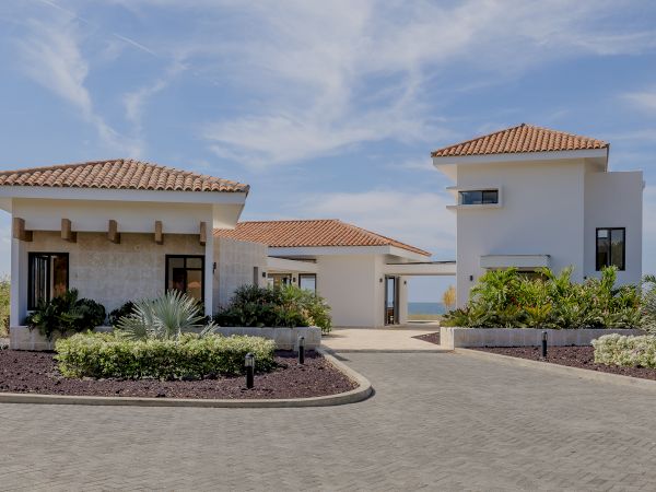 The image shows a modern villa complex with two buildings, paved pathways, landscaped gardens, and a clear blue sky.