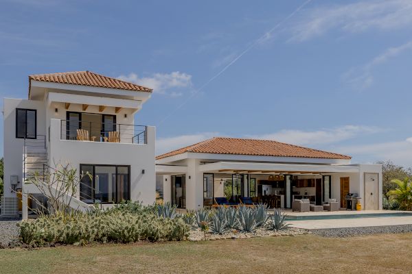 A modern two-story house with a pool, outdoor dining area, and plants in the yard is shown under a clear blue sky.