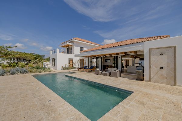 A modern home with a swimming pool, outdoor seating area, and lush vegetation in the background, under a clear blue sky.