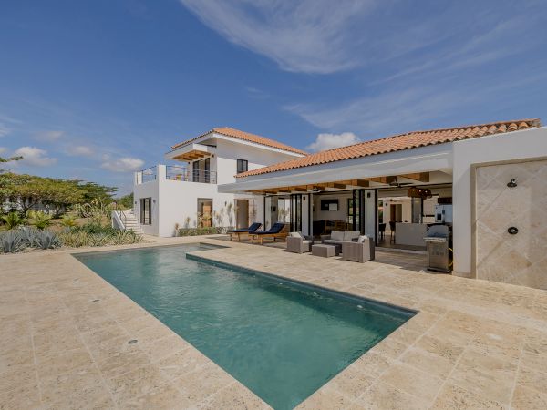A modern home with a swimming pool, outdoor seating area, and lush vegetation in the background, under a clear blue sky.
