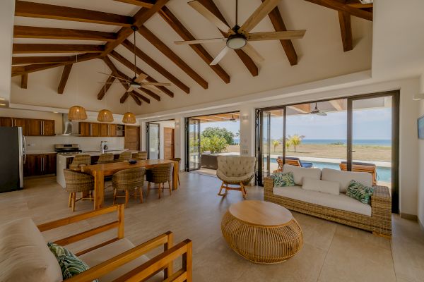 The image shows a modern, open-plan living area with high ceilings, wood beams, and large windows offering a view of a pool and the ocean.