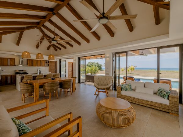 The image shows a modern, open-plan living area with high ceilings, wood beams, and large windows offering a view of a pool and the ocean.