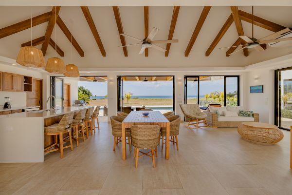 Open-concept living space with wooden beams, dining area, and kitchen; large windows with ocean view and outdoor patio.