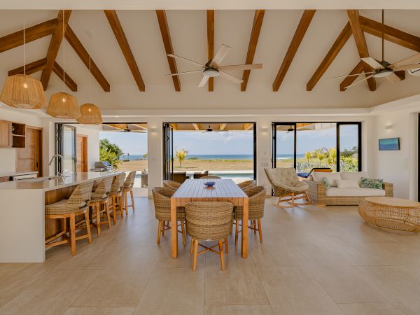 Open-concept living space with wooden beams, dining area, and kitchen; large windows with ocean view and outdoor patio.