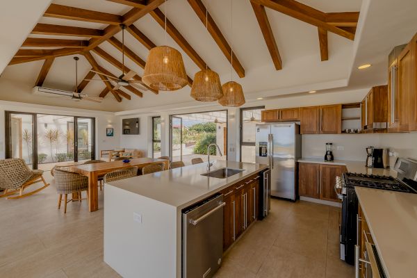 A modern kitchen with wooden cabinets, an island, stainless steel appliances, and hanging lights, leading to a dining area with large windows.