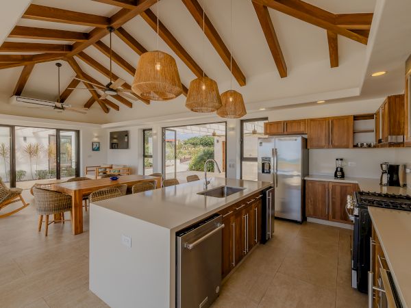 A modern kitchen with wooden cabinets, an island, stainless steel appliances, and hanging lights, leading to a dining area with large windows.