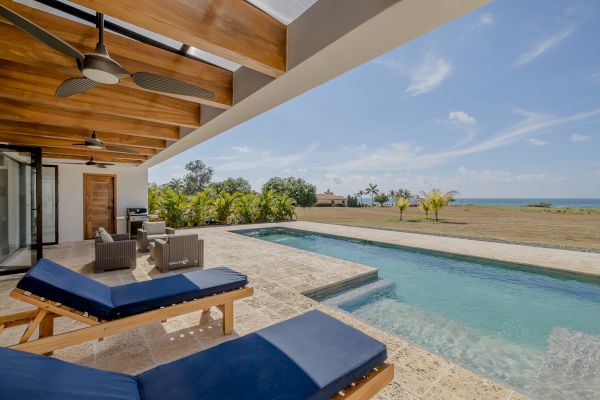 An outdoor patio features lounge chairs, a pool, and a covered seating area with ceiling fans, overlooking a spacious lawn and ocean in the distance.