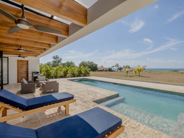 An outdoor patio features lounge chairs, a pool, and a covered seating area with ceiling fans, overlooking a spacious lawn and ocean in the distance.